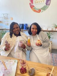Two smiling women in aprons holding homemade natural skincare jars and bottles at a bright DIY skincare workshop, with jars of dried rose petals and botanicals on the table.