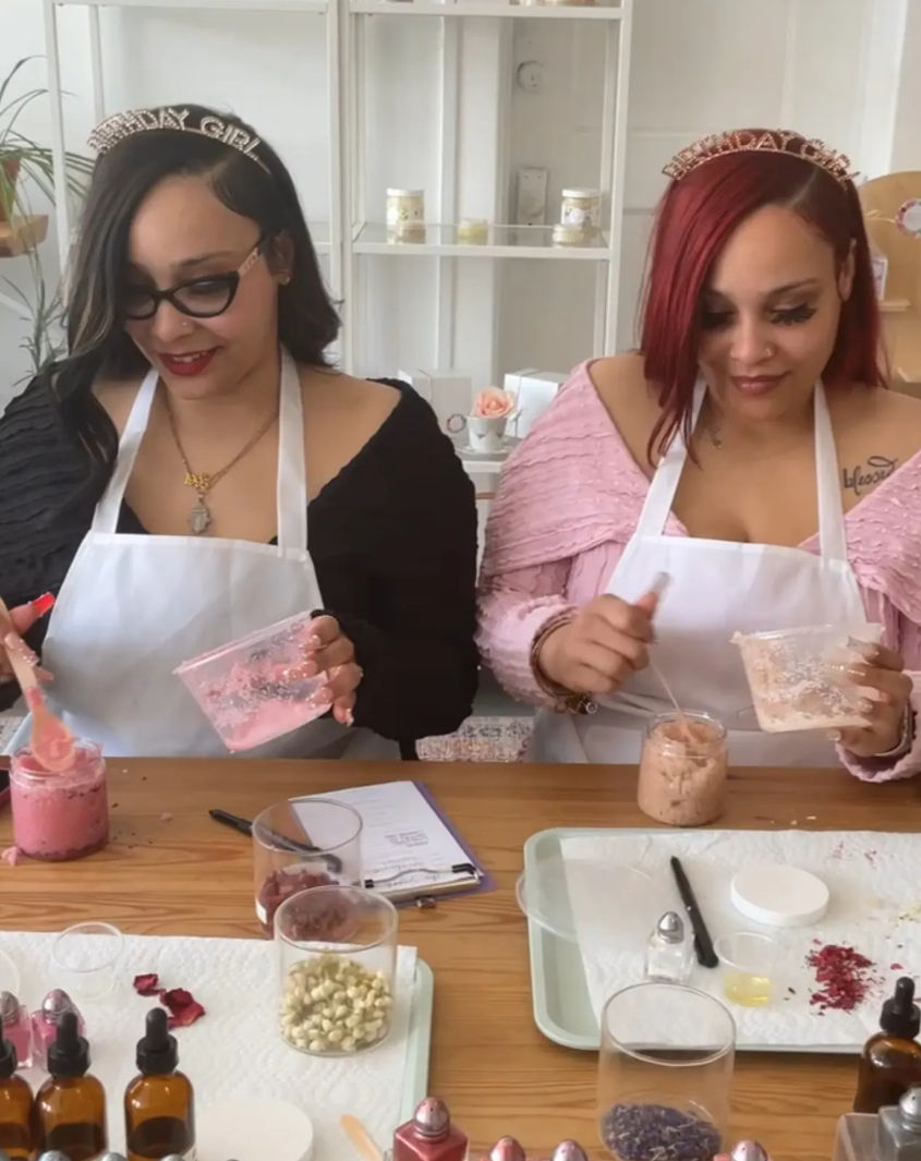Two people in aprons and "Birthday Girl" tiaras stirring pink mixtures at a wooden table in a DIY bath-scrub and skincare workshop, surrounded by jars of botanicals, oils, droppers and mixing tools.