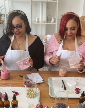 Two people in aprons and "Birthday Girl" tiaras stirring pink mixtures at a wooden table in a DIY bath-scrub and skincare workshop, surrounded by jars of botanicals, oils, droppers and mixing tools.
