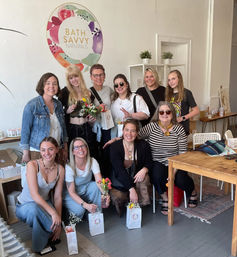 Smiling group of women posing in a bright boutique studio, holding gift bags and fresh flower bouquets under colorful circular wall art.