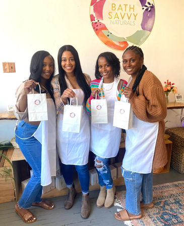 Four women in aprons smiling and holding white gift bags inside a bright bath-and-body studio with a pastel circular wall sign, wooden shelves and plants — DIY bath products workshop vibe