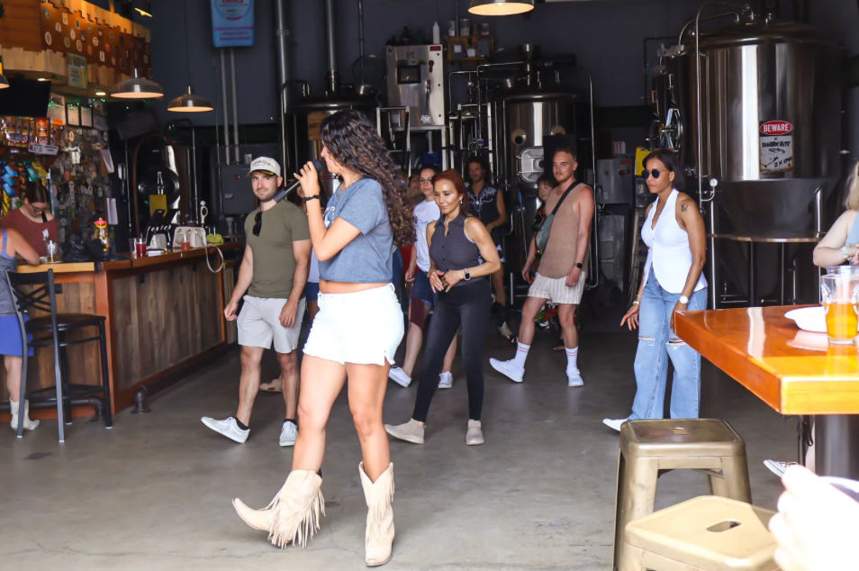 People dancing and socializing in a microbrewery taproom with stainless steel fermenters; a woman in fringe cowboy boots sings into a microphone near the bar.