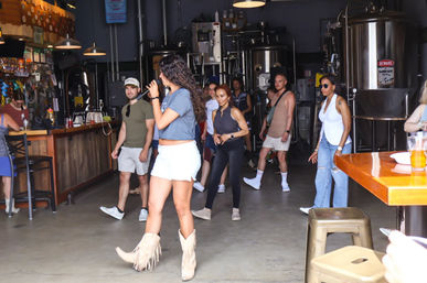 People dancing and socializing in a microbrewery taproom with stainless steel fermenters; a woman in fringe cowboy boots sings into a microphone near the bar.