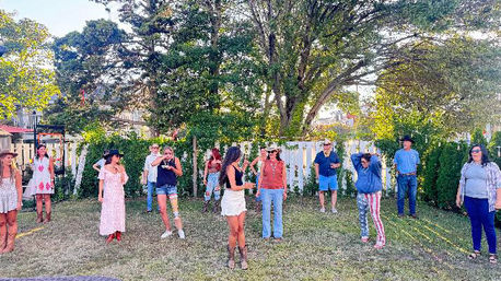 Backyard summer gathering on a grassy lawn by a white picket fence and large shade tree, a group of casually dressed people wearing boots, hats and summer outfits spread out and chatting.