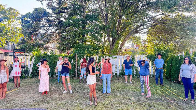 Backyard summer gathering on a grassy lawn by a white picket fence and large shade tree, a group of casually dressed people wearing boots, hats and summer outfits spread out and chatting.