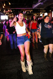 Young woman in a white crop top and denim shorts leading a lively line dance on a crowded indoor bar dance floor, wearing fringed cowboy boots and pointing toward the camera under string lights.