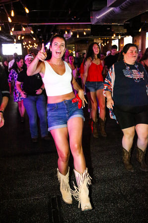 Young woman in a white crop top and denim shorts leading a lively line dance on a crowded indoor bar dance floor, wearing fringed cowboy boots and pointing toward the camera under string lights.