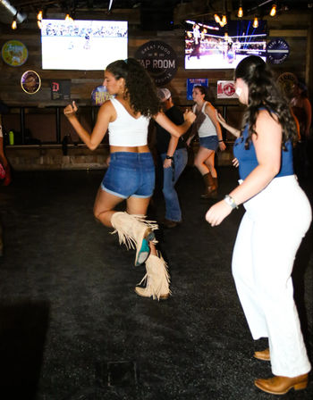 People line-dancing on a rustic indoor bar dance floor — a woman in fringe tan cowboy boots and denim shorts kicks mid-step beneath wall-mounted TVs and warm pendant lights.