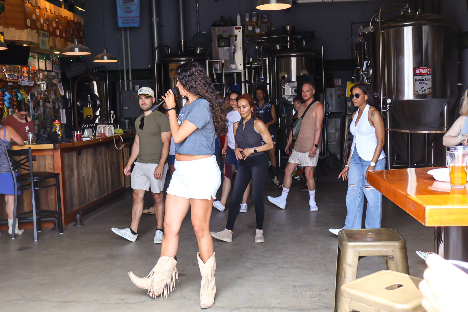Woman in fringe cowboy boots and white shorts leading a small crowd dancing and singing inside a craft brewery taproom with stainless steel brewing tanks and a wooden bar.