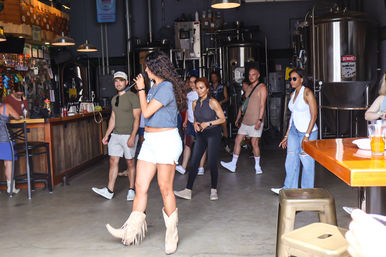 Woman in fringe cowboy boots and white shorts leading a small crowd dancing and singing inside a craft brewery taproom with stainless steel brewing tanks and a wooden bar.