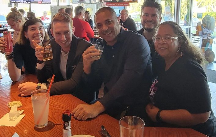 Five friends smiling at a casual restaurant bar, seated around a wooden table holding cocktails and cans during happy hour with large windows and a lively indoor dining background.