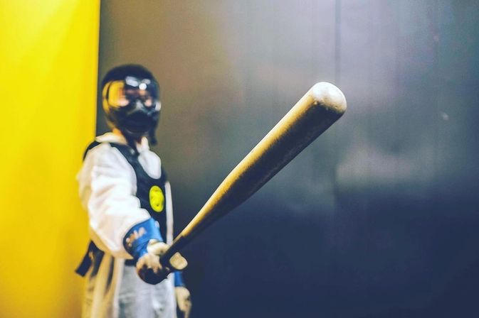 Masked figure in white protective suit and goggles pointing a weathered baseball bat toward the camera against a yellow and dark indoor background