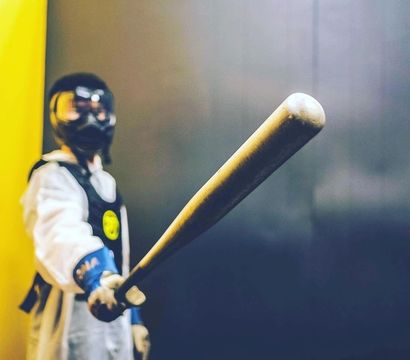 Masked figure in white protective suit and goggles pointing a weathered baseball bat toward the camera against a yellow and dark indoor background