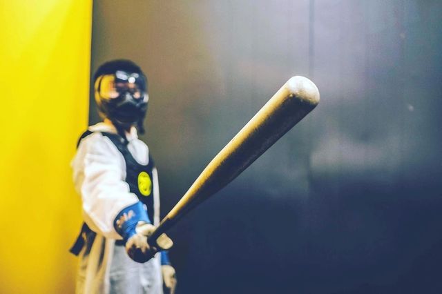 Masked figure in white protective suit and goggles pointing a weathered baseball bat toward the camera against a yellow and dark indoor background
