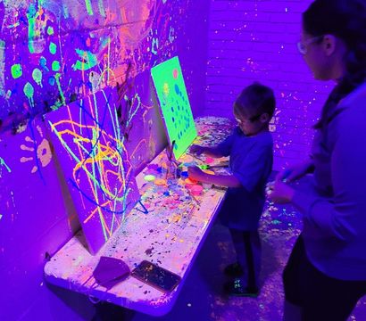 Child making neon UV paintings at an indoor blacklight art studio, glowing canvases and splatter-covered table and walls under purple light