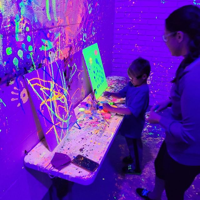 Child making neon UV paintings at an indoor blacklight art studio, glowing canvases and splatter-covered table and walls under purple light