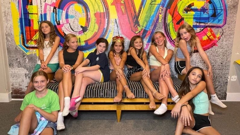 Group of girls posing on a striped bench in an indoor lounge, smiling in casual summer outfits and socks in front of a vibrant multicolored "LOVE" mural.