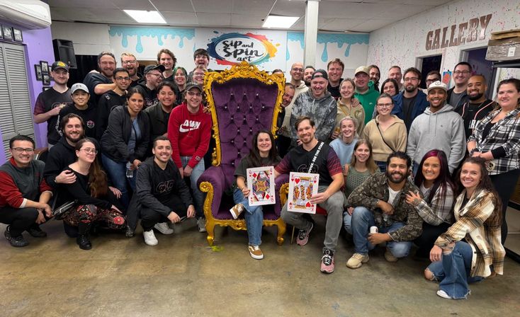 Large group photo in a colorful paint-and-sip studio gathered around an ornate purple-and-gold throne, two people seated holding oversized playing cards, smiling near a gallery wall.
