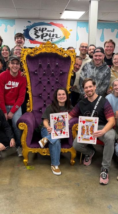 Large group photo in a colorful paint-and-sip studio gathered around an ornate purple-and-gold throne, two people seated holding oversized playing cards, smiling near a gallery wall.