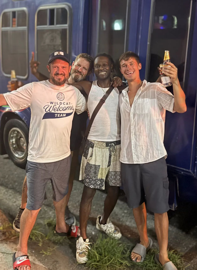 Four smiling friends with arms around each other, holding beer bottles and posing in casual summer clothes on a nighttime street in front of a blue bus — fun night out.