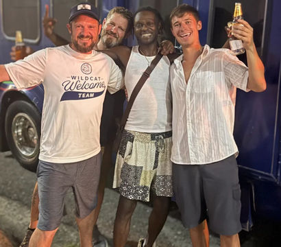 Four smiling friends with arms around each other, holding beer bottles and posing in casual summer clothes on a nighttime street in front of a blue bus — fun night out.