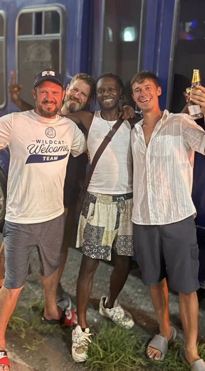 Four smiling friends with arms around each other, holding beer bottles and posing in casual summer clothes on a nighttime street in front of a blue bus — fun night out.