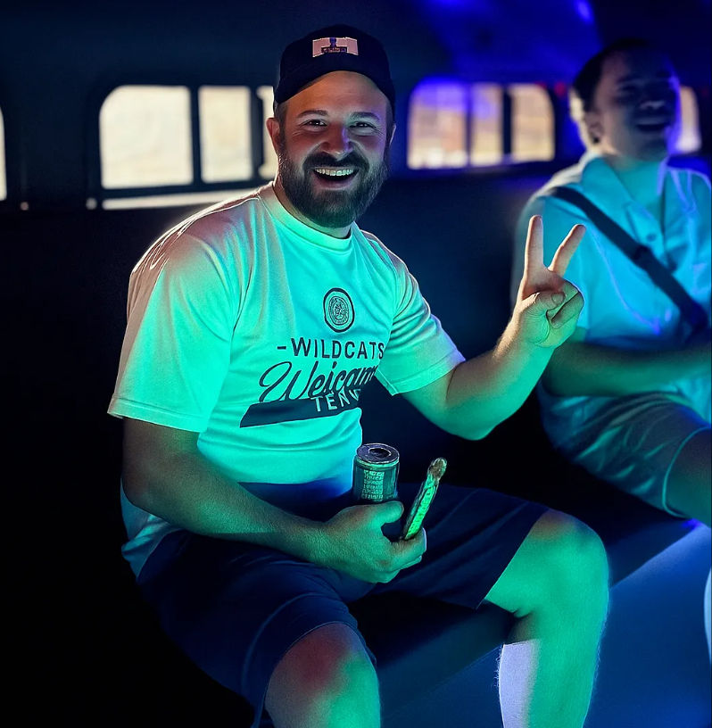 Smiling bearded man in a cap flashes a peace sign inside a neon-lit party vehicle, wearing a 'Wildcats Welcome' tee and holding a canned drink and snack, with a laughing friend blurred in the background.