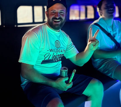 Smiling bearded man in a cap flashes a peace sign inside a neon-lit party vehicle, wearing a 'Wildcats Welcome' tee and holding a canned drink and snack, with a laughing friend blurred in the background.