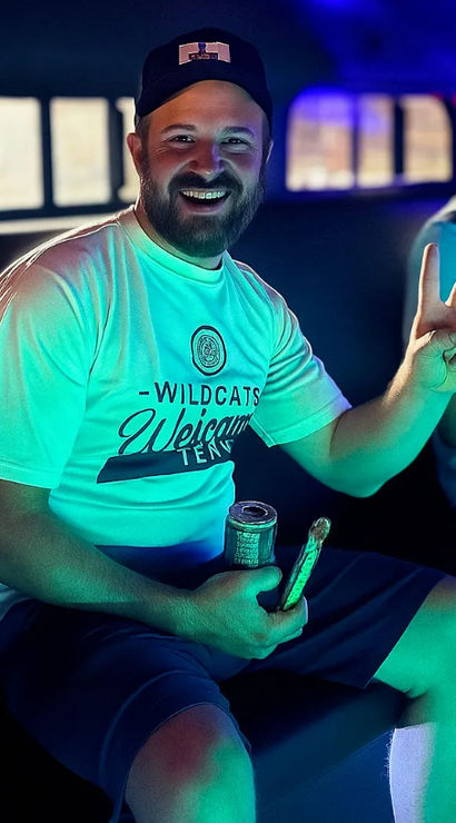 Smiling bearded man in a cap flashes a peace sign inside a neon-lit party vehicle, wearing a 'Wildcats Welcome' tee and holding a canned drink and snack, with a laughing friend blurred in the background.