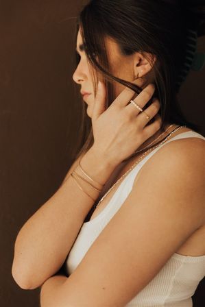 Side profile of a person in a white ribbed tank touching their hair, showcasing delicate gold bracelets, a pearl beaded ring, thin stacked rings and layered necklaces against a warm brown background.
