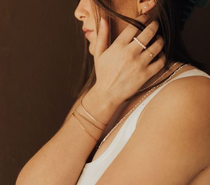 Side profile of a person in a white ribbed tank touching their hair, showcasing delicate gold bracelets, a pearl beaded ring, thin stacked rings and layered necklaces against a warm brown background.
