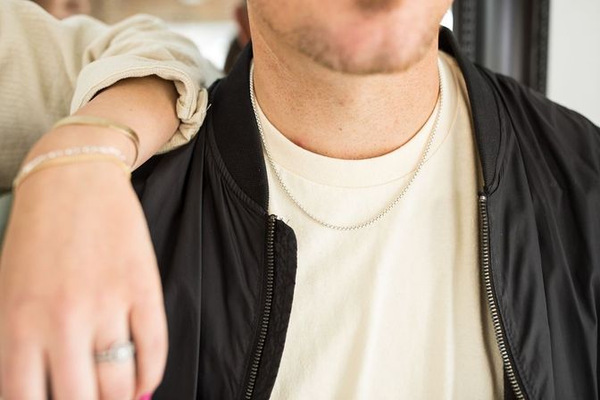 Close-up of two people: one wearing a cream T‑shirt and black bomber jacket with layered silver chain necklaces, another's arm draped over the shoulder showing bracelets and a ring — casual jewelry and street-style outfit.