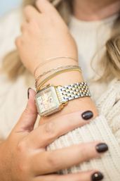 Close-up of a wrist wearing a two-tone silver-and-gold square watch with a diamond bezel, layered delicate gold and silver bracelets, dark polished nails, and a cream knit sweater — stylish watch and bracelet stack.