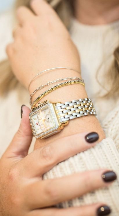 Close-up of a wrist wearing a two-tone silver-and-gold square watch with a diamond bezel, layered delicate gold and silver bracelets, dark polished nails, and a cream knit sweater — stylish watch and bracelet stack.