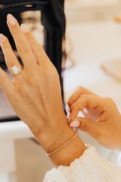 Close-up of a manicured hand with pale pink nails, showcasing a sparkling cushion-cut diamond engagement ring while adjusting two delicate gold bracelets — layered jewelry detail.