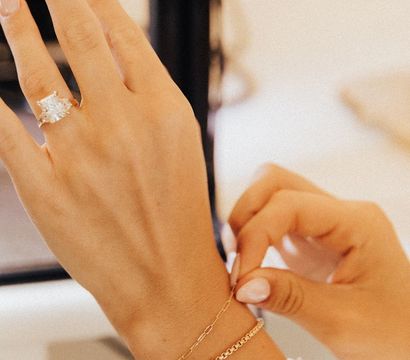 Close-up of a manicured hand with pale pink nails, showcasing a sparkling cushion-cut diamond engagement ring while adjusting two delicate gold bracelets — layered jewelry detail.