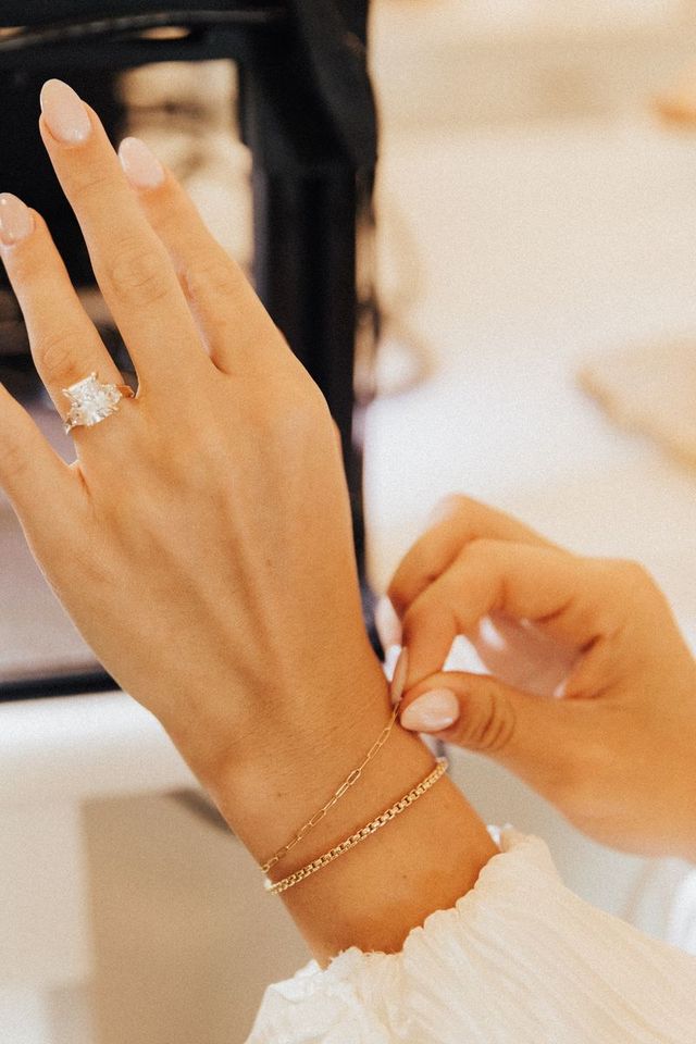 Close-up of a manicured hand with pale pink nails, showcasing a sparkling cushion-cut diamond engagement ring while adjusting two delicate gold bracelets — layered jewelry detail.