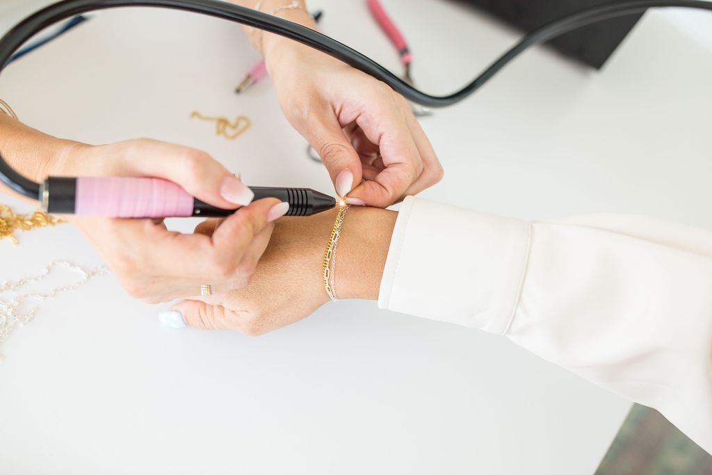 Close-up of hands using a pink-handled rotary tool to repair a delicate gold bracelet on a wrist at a white jewelry studio workbench.