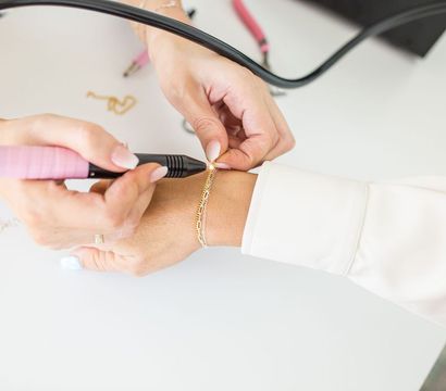 Close-up of hands using a pink-handled rotary tool to repair a delicate gold bracelet on a wrist at a white jewelry studio workbench.