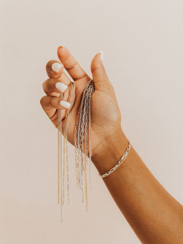 Close-up of a manicured hand holding cascading gold and silver chain necklaces with a delicate bracelet against a neutral beige background