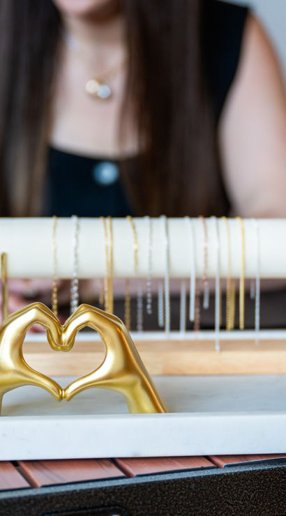 Close-up of a tabletop jewelry display with a marble tray holding a gold hands-formed heart sculpture and delicate gold and silver necklaces on a stand, blurred person working in the background in a boutique studio.