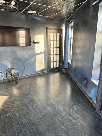 Empty dark-blue interior with scuffed black hardwood floor, wall-mounted flat-screen TV, metal floor fan, glass-paneled door and sunlight streaming through a tall window
