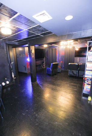 Dim basement entertainment/game room with glossy black floor and tiled ceiling, recliner seats facing wall TVs, dark curtains and a shelf of board games.