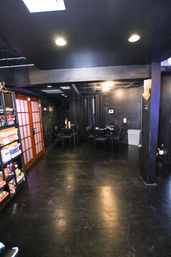Dim black-painted game room with a shelf of board games beside wooden sliding frosted doors, small tables and chairs under recessed ceiling lights, curtains and a trash bin — ready for game night.