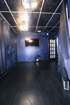 Moody interior studio with dark blue walls, black hardwood floor, wall-mounted flat-screen TV, industrial cage ceiling lights, black curtains and a floor fan.