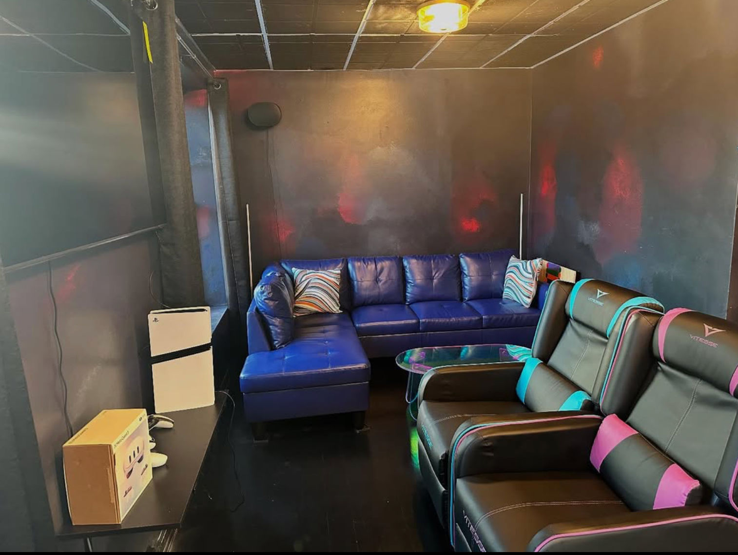 Cozy, neon-lit home gaming lounge with a blue leather sectional, two black gaming recliners with teal and pink accents, glass coffee table, large flat-screen TV and dark walls with colorful ambient lighting.