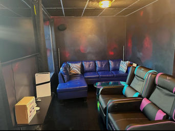 Cozy, neon-lit home gaming lounge with a blue leather sectional, two black gaming recliners with teal and pink accents, glass coffee table, large flat-screen TV and dark walls with colorful ambient lighting.