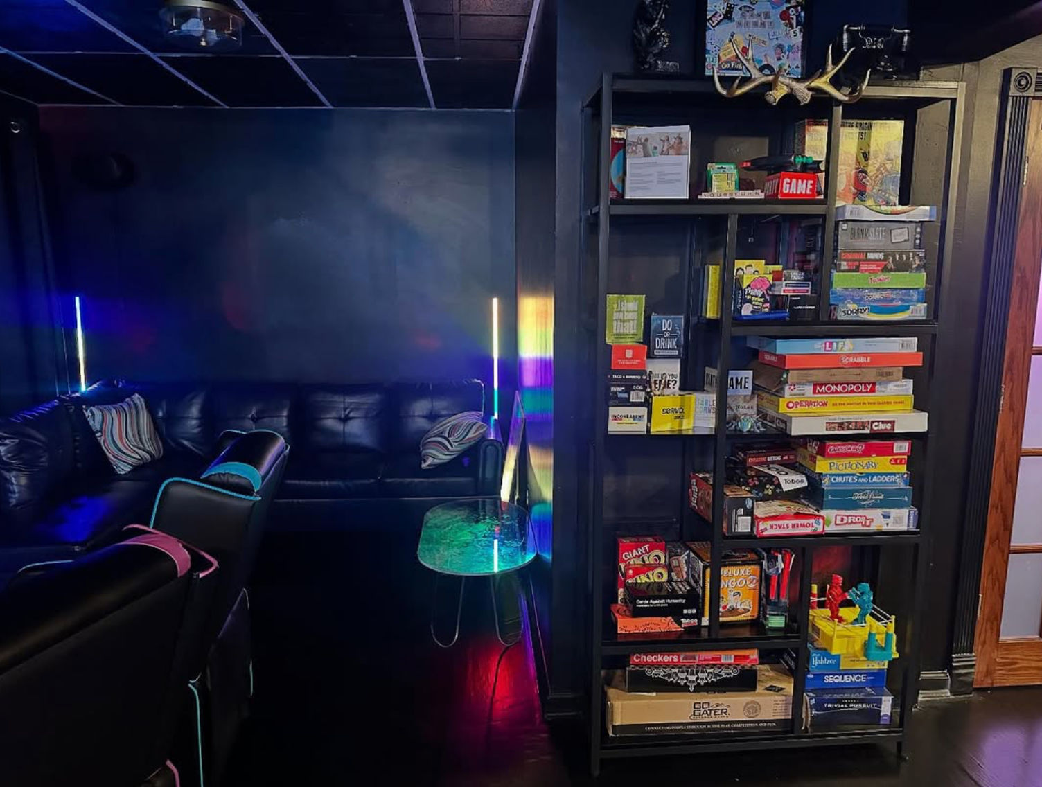 Cozy dark game room with black leather sectional and recliners, tall shelving stacked with board games, neon RGB floor lights casting colorful reflections, small glass side table and striped throw pillows.
