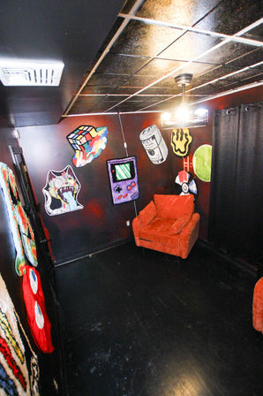 Small eclectic entertainment room interior with a bright orange armchair against dark red walls, black tiled ceiling and floor, pendant light overhead, and colorful pop-art wall rugs including a Game Boy, Rubik’s Cube, Poké Ball, rolled cash and whimsical monster motifs.