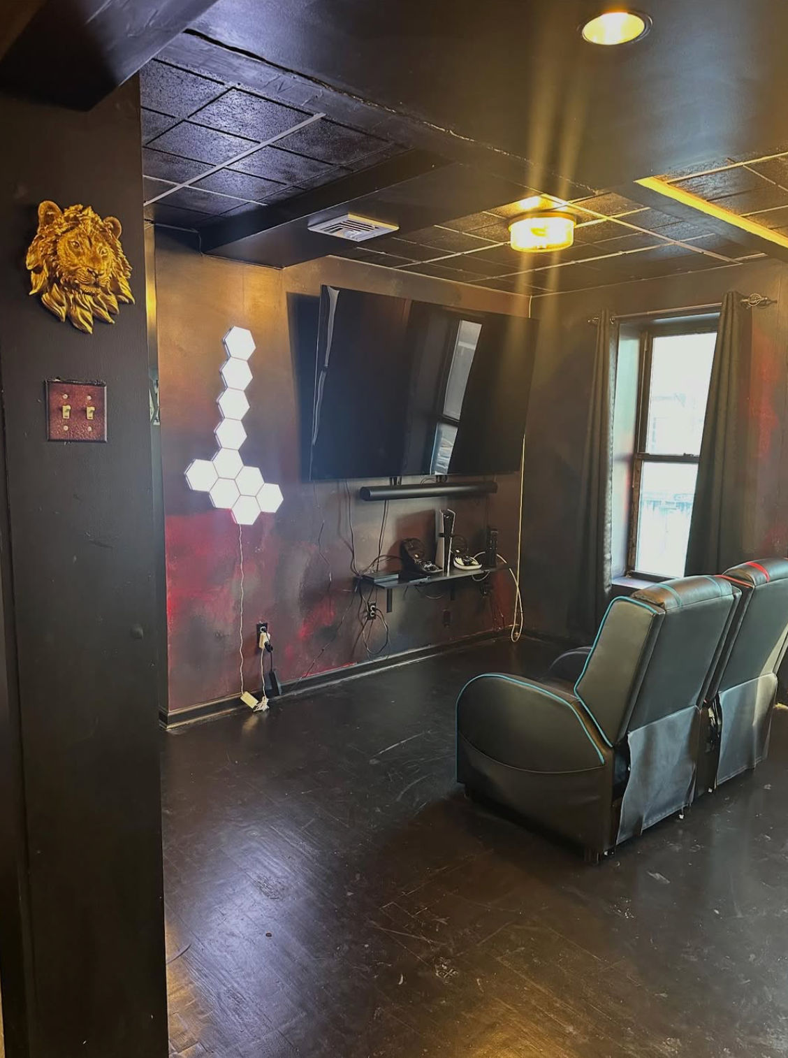 Dark home theater-style room with wall-mounted flat-screen TV, two black leather recliners, hexagonal LED wall lights, lion head decor, recessed ceiling lights and a curtained window.